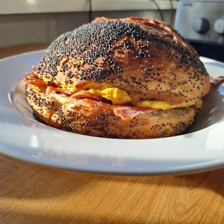 A close-up photo shows a large, round sandwich with a poppy seed bun, filled with scrambled eggs and bacon, sitting on a…