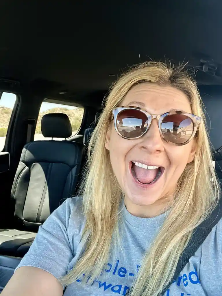 A woman with blonde hair and sunglasses is taking a selfie inside a car, sticking her tongue out playfully while wearing a…