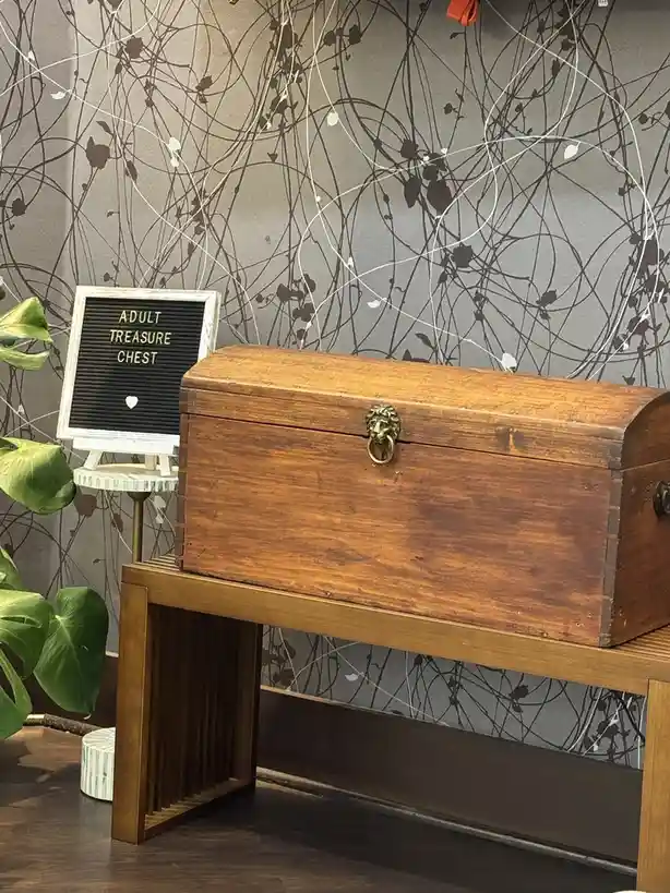 A wooden treasure chest with a lion-head knob sits on a wooden bench next to a small white stand holding a black letterboard…