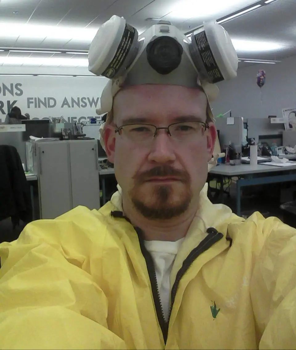 A man in a yellow hazmat-style jacket and glasses wears a full-face gas mask on his head, looking directly at the camera…