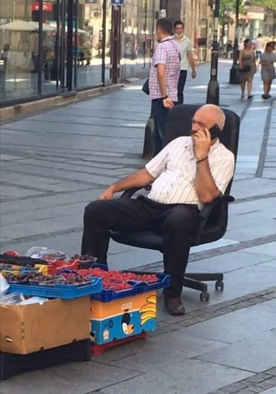 A man sits in a large office chair on a city sidewalk next to crates of fruit, talking on a cell phone. He appears relaxed…