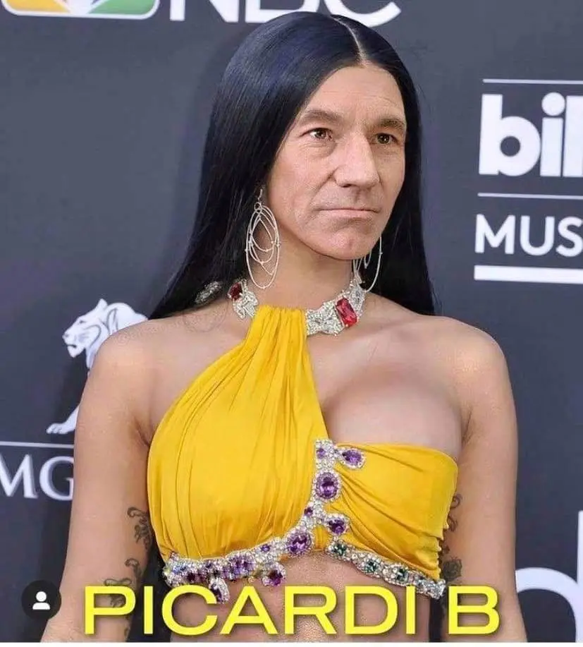 A man with long black hair and a serious expression is dressed in a glamorous yellow one-shoulder outfit adorned with large…