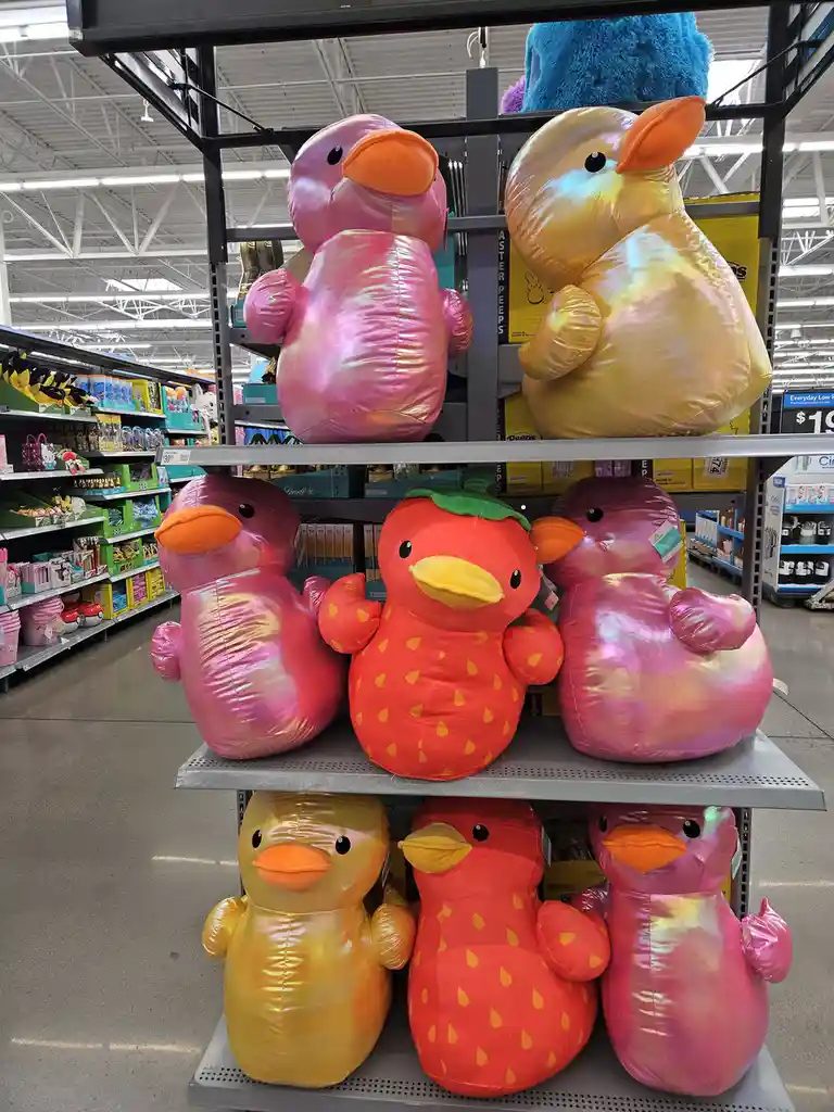 A photo of large, shiny plush ducks in a store aisle, with one duck shaped like a strawberry and wearing a green leaf on its…
