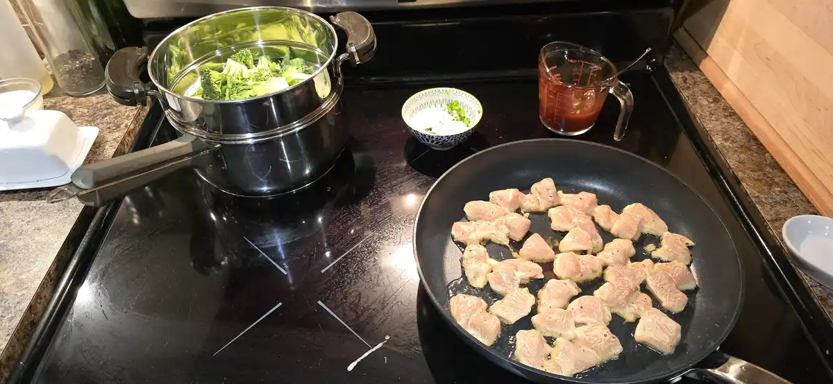 A kitchen scene shows a stovetop with a frying pan full of chicken pieces sizzling and a pot of broccoli steaming. Nearby…