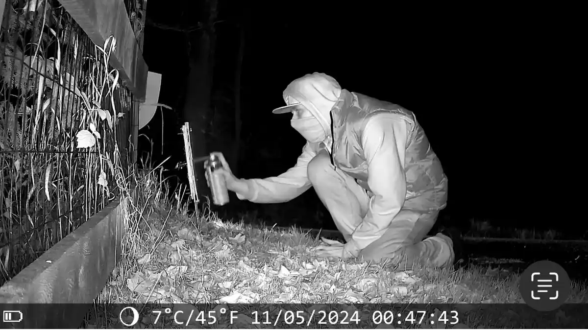 A black-and-white night-vision security camera image shows a person in a hooded jacket and face mask crouching beside a…