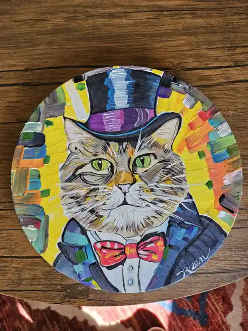 A hand-painted portrait on a circular canvas depicts a fluffy cat with green eyes wearing a black top hat with a purple…