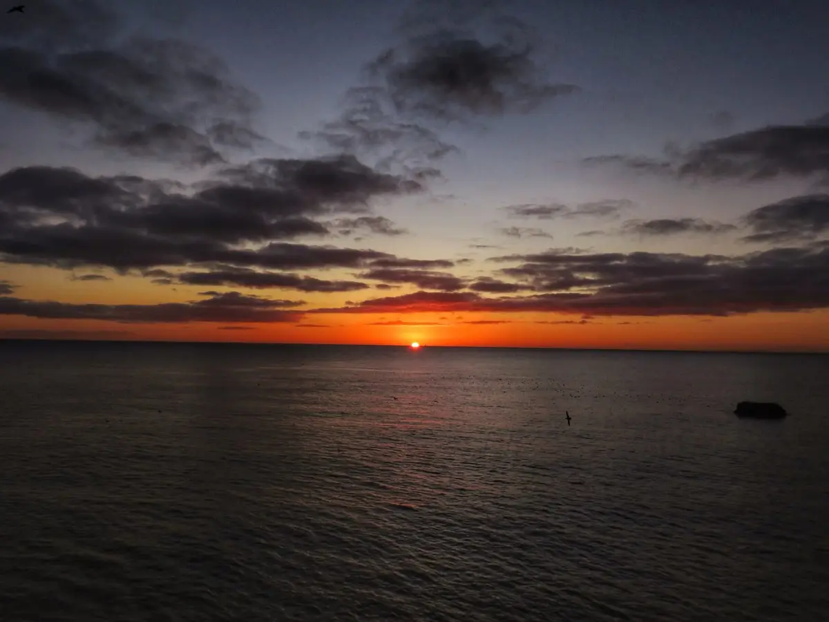 A serene sunset over the ocean with the sun low on the horizon, casting orange and purple hues across the sky and water…