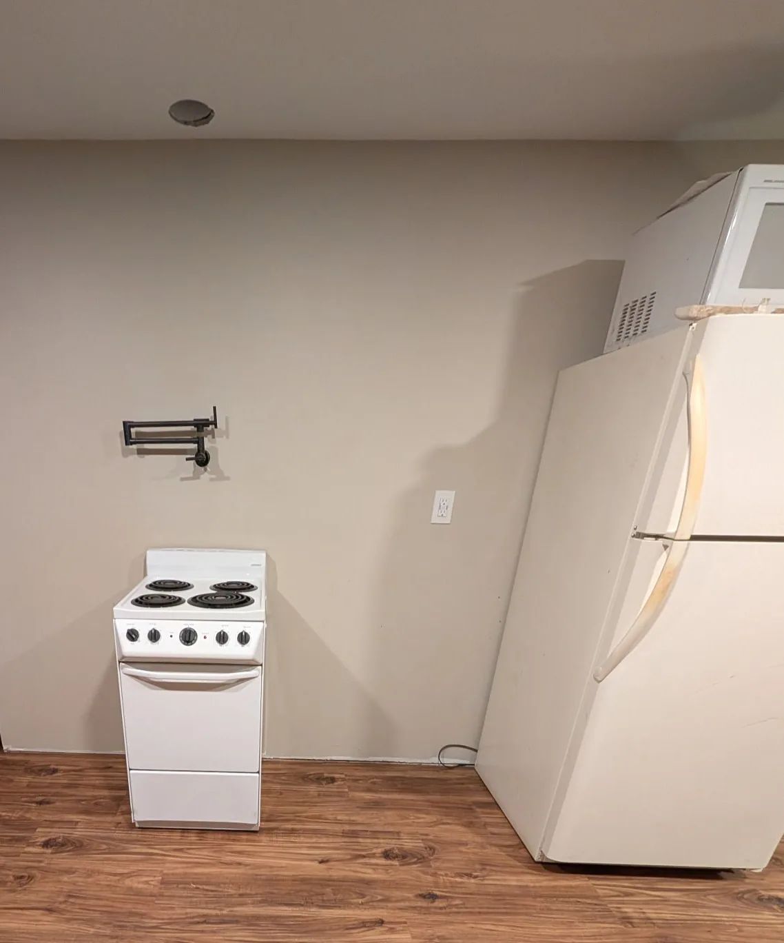 A sparse kitchen with a white electric stove and a white refrigerator, both slightly off-center, on wood-patterned flooring…