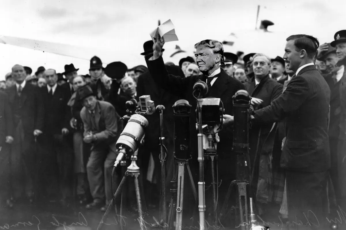 a black-and-white historical photo shows a man in a suit holding up a piece of paper while surrounded by vintage cameras on…