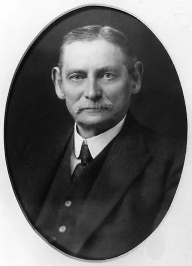 A vintage black-and-white oval portrait of a stern-looking man with a mustache, wearing a suit and tie, overlaid with the…