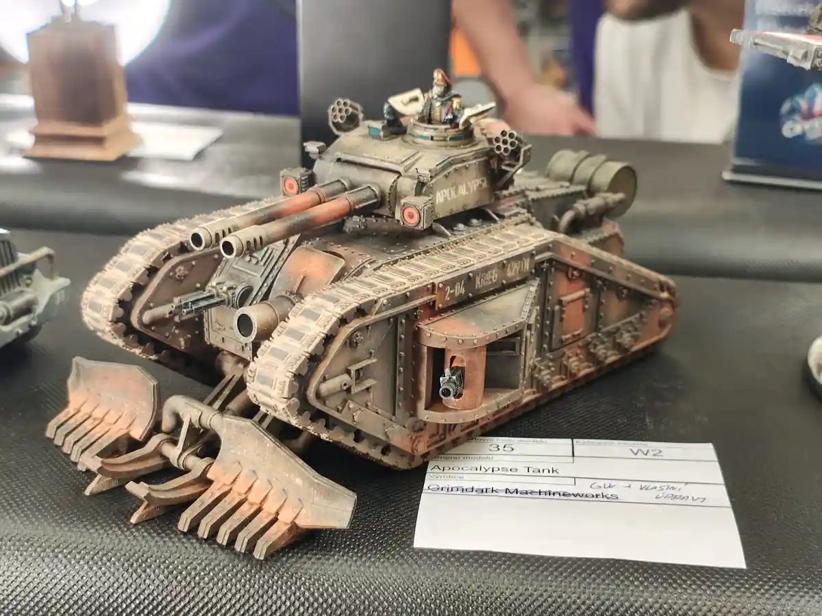 A highly detailed, weathered miniature Apocalypse Tank model from Warhammer 40,000 sits on a display table with a label…