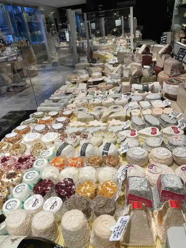 A wide variety of cheeses are displayed in a shop, arranged in rows on wooden trays, with some wrapped in plastic and others…