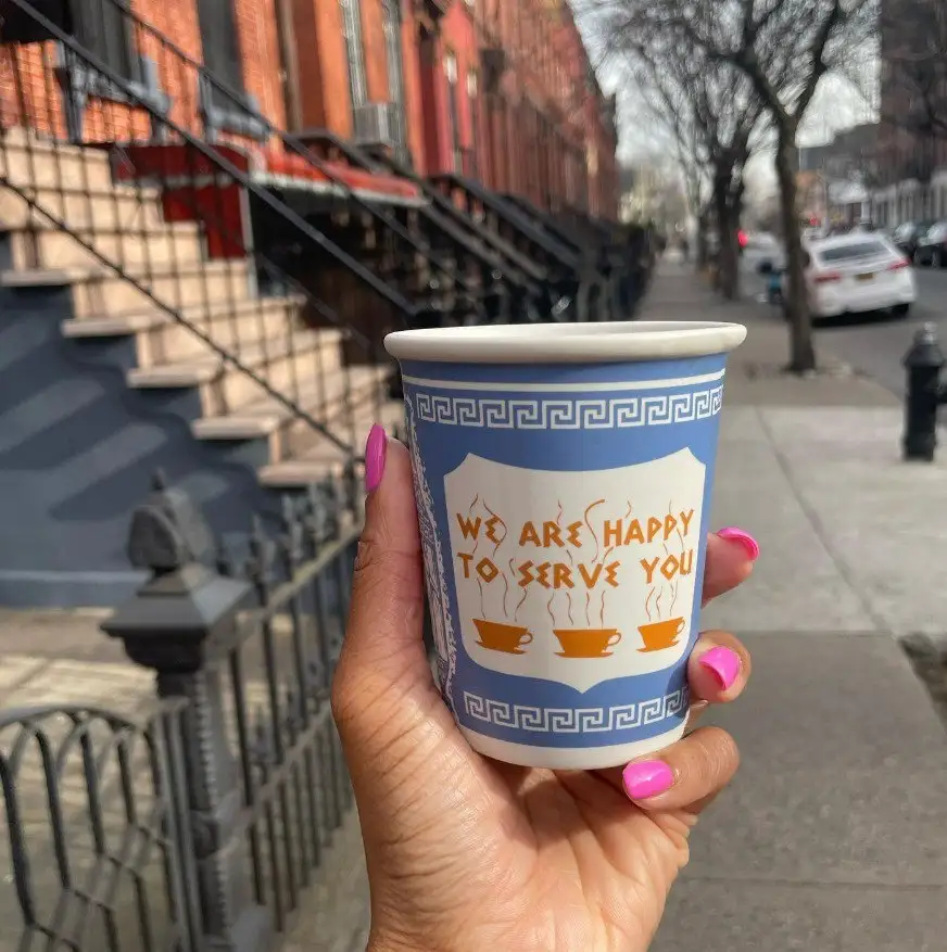 A hand with bright pink fingernails holds a blue paper coffee cup with a white shield design and Greek key border. The cup…
