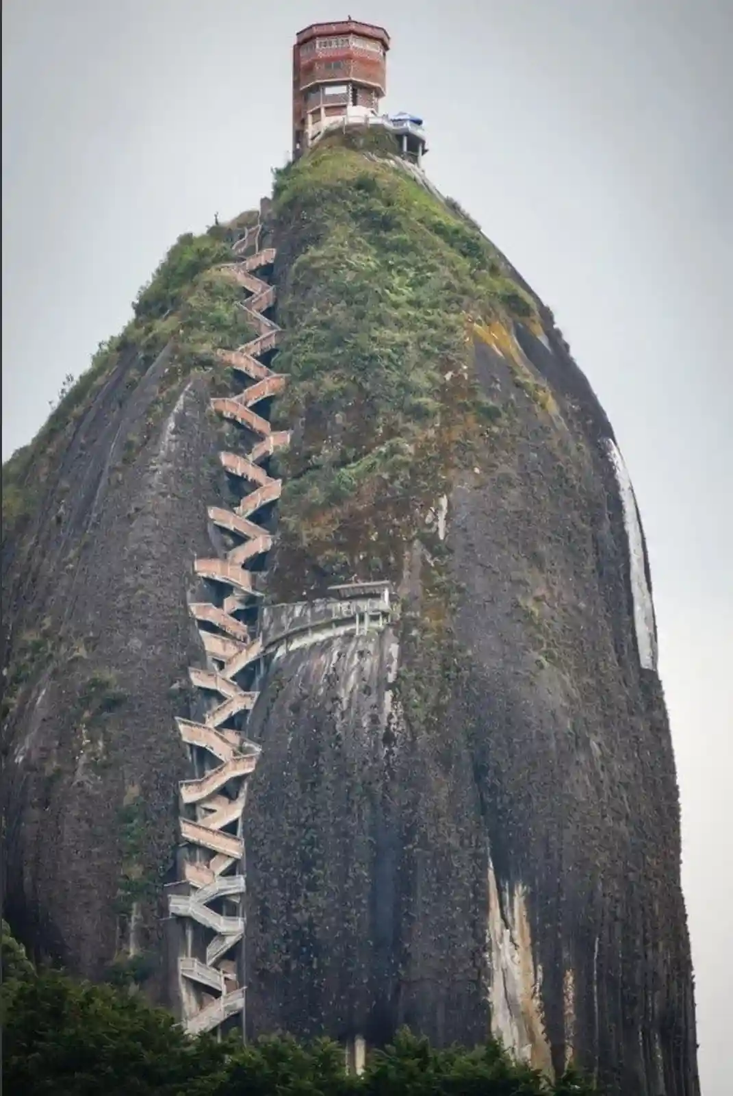 A steep, dark rock mountain with a winding staircase zigzagging up its side to a small building at the peak. The stairs are…