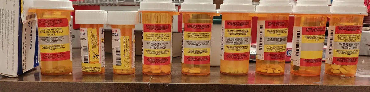 A row of orange prescription pill bottles with white caps sits on a countertop, each displaying different medication labels…