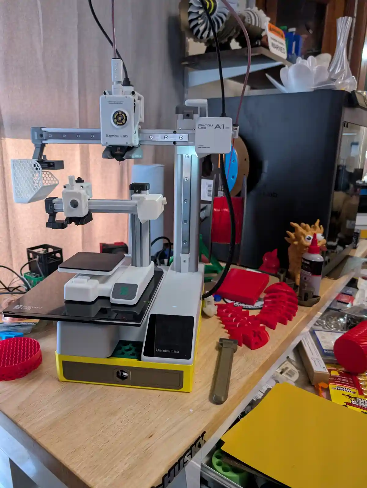 A Bambu Lab A1 3D printer sits on a cluttered wooden workbench, surrounded by red 3D-printed objects, spools of filament…