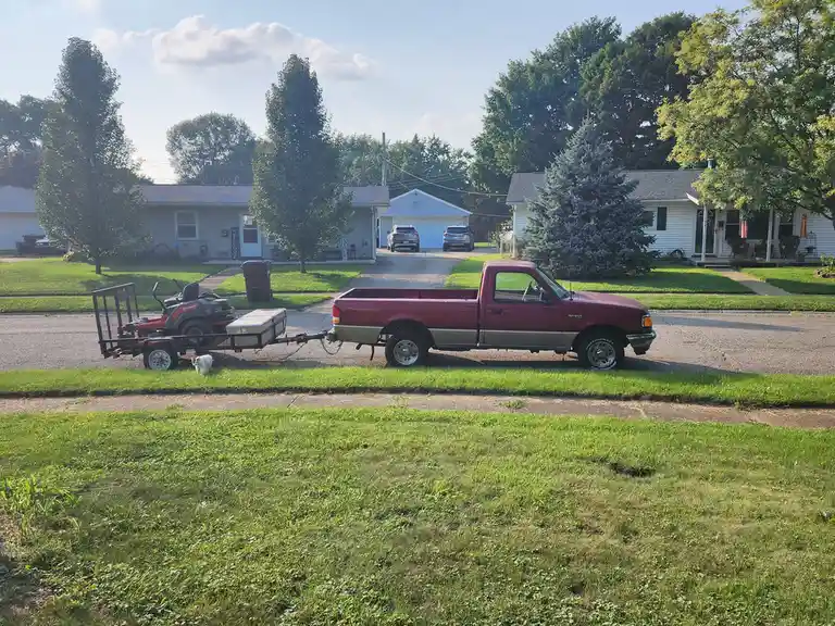 A maroon pickup truck is parked on a residential street, hitched to a trailer carrying a riding lawnmower and a storage box…
