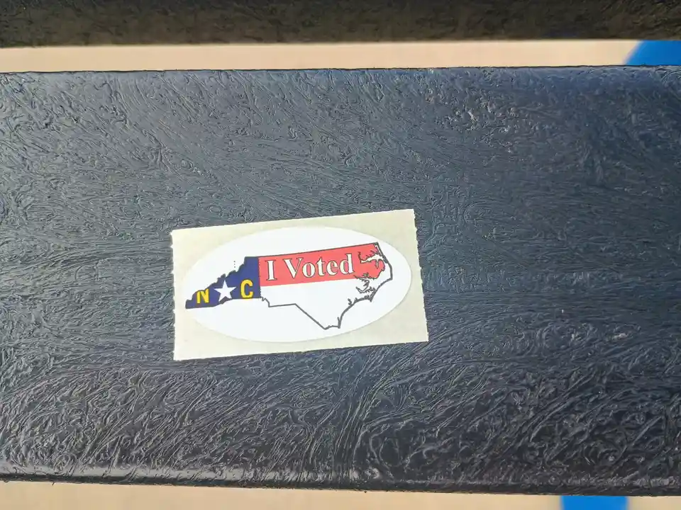 A North Carolina 'I Voted' sticker is stuck on a dark, textured surface, likely a bench or railing. The sticker features the…