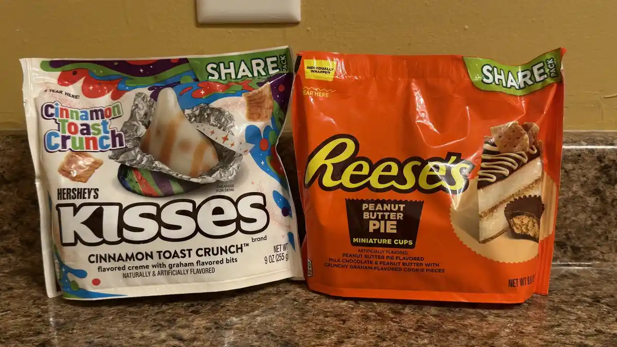 Two bags of candy sit side by side on a countertop: a white bag of Hershey's Kisses in Cinnamon Toast Crunch flavor and an…