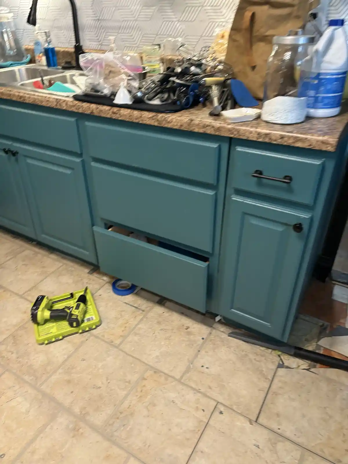 A messy kitchen counter with teal cabinets, one drawer partially open, and a green power drill on the tiled floor. The…