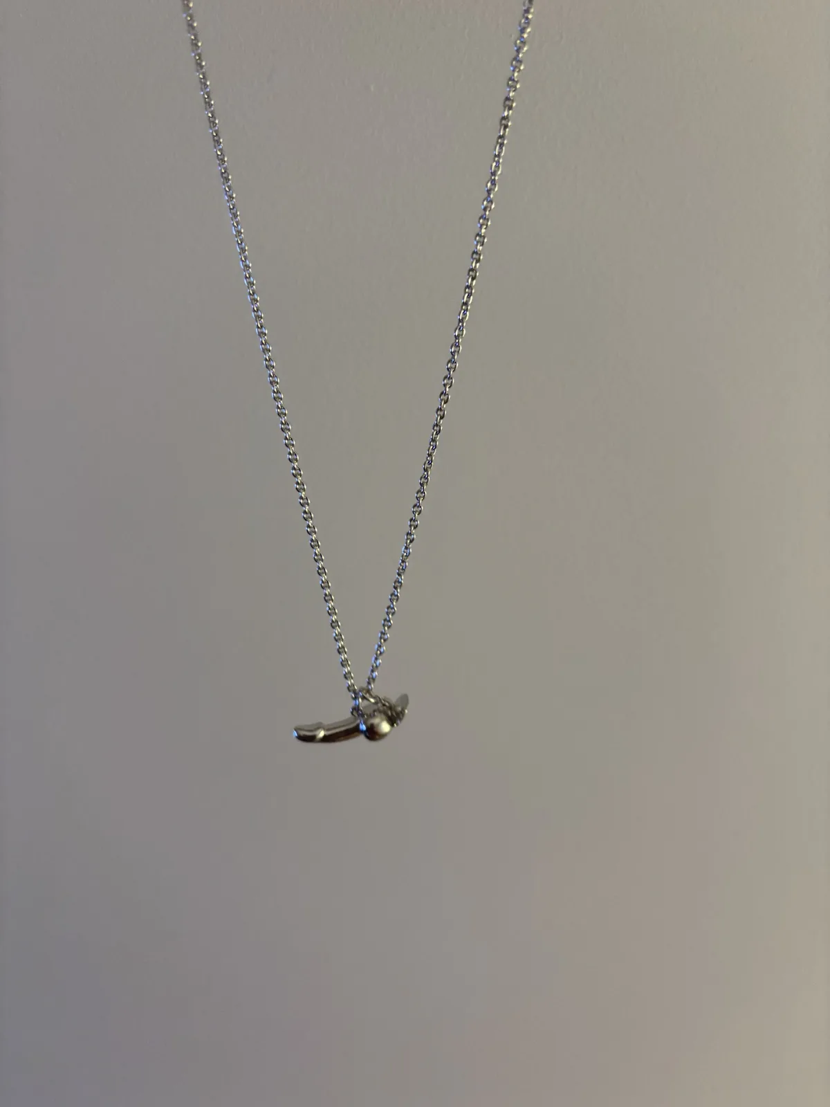 A silver chain necklace hangs against a plain light gray wall, ending in a small, curved pendant shaped like a vintage…