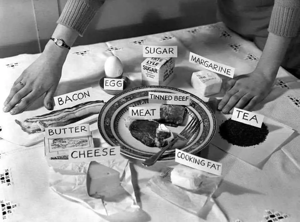 A black-and-white photo shows a person's hands resting on a table covered with various food items, each labeled with a white…