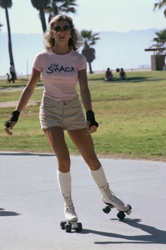A woman in 1980s-style attire, including a pink t-shirt with 'the Shack' printed on it, beige shorts, white roller skates…