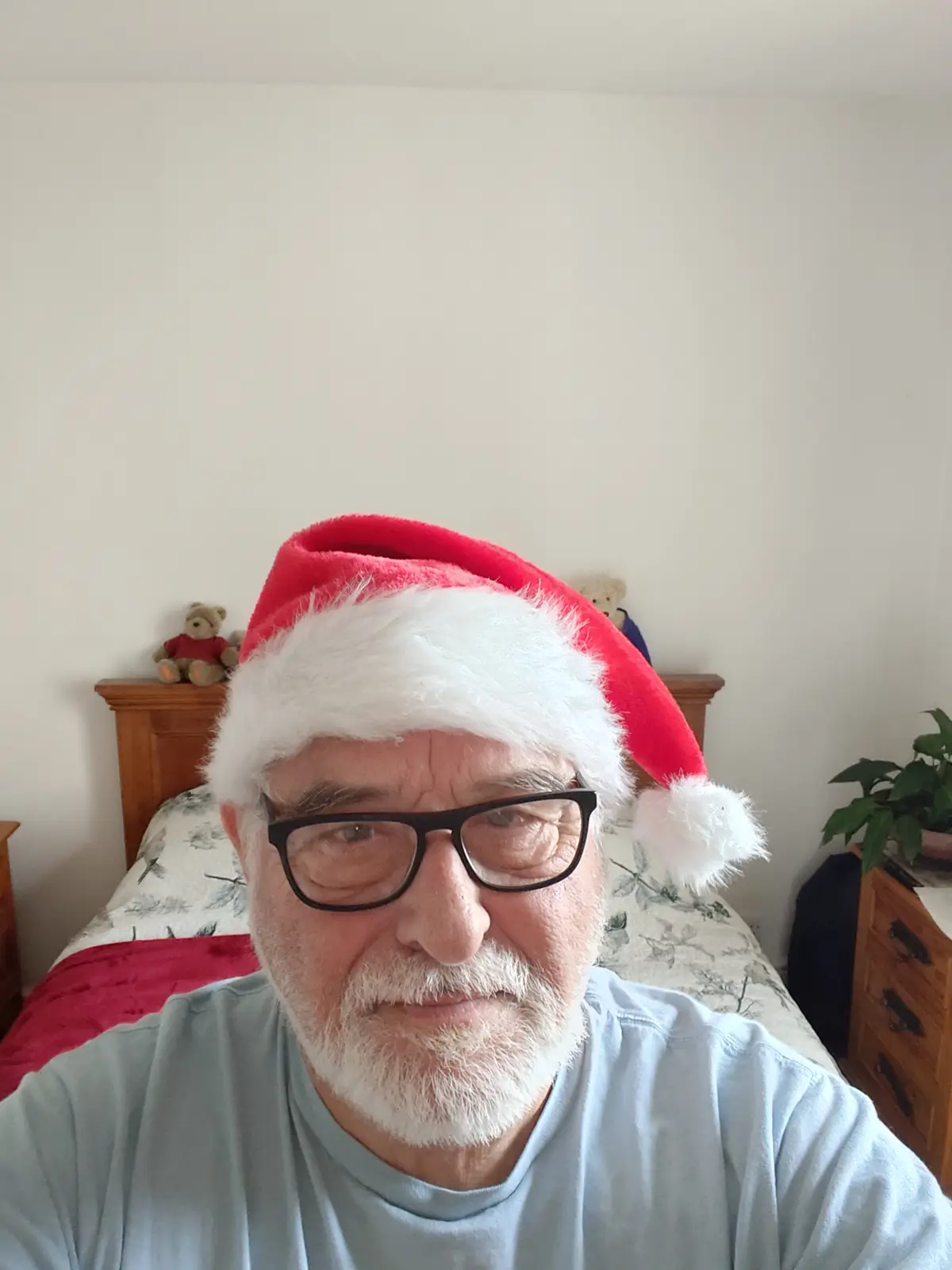 An older man with a white beard and glasses wears a red and white Santa hat while sitting on a bed in a bedroom. Behind him…