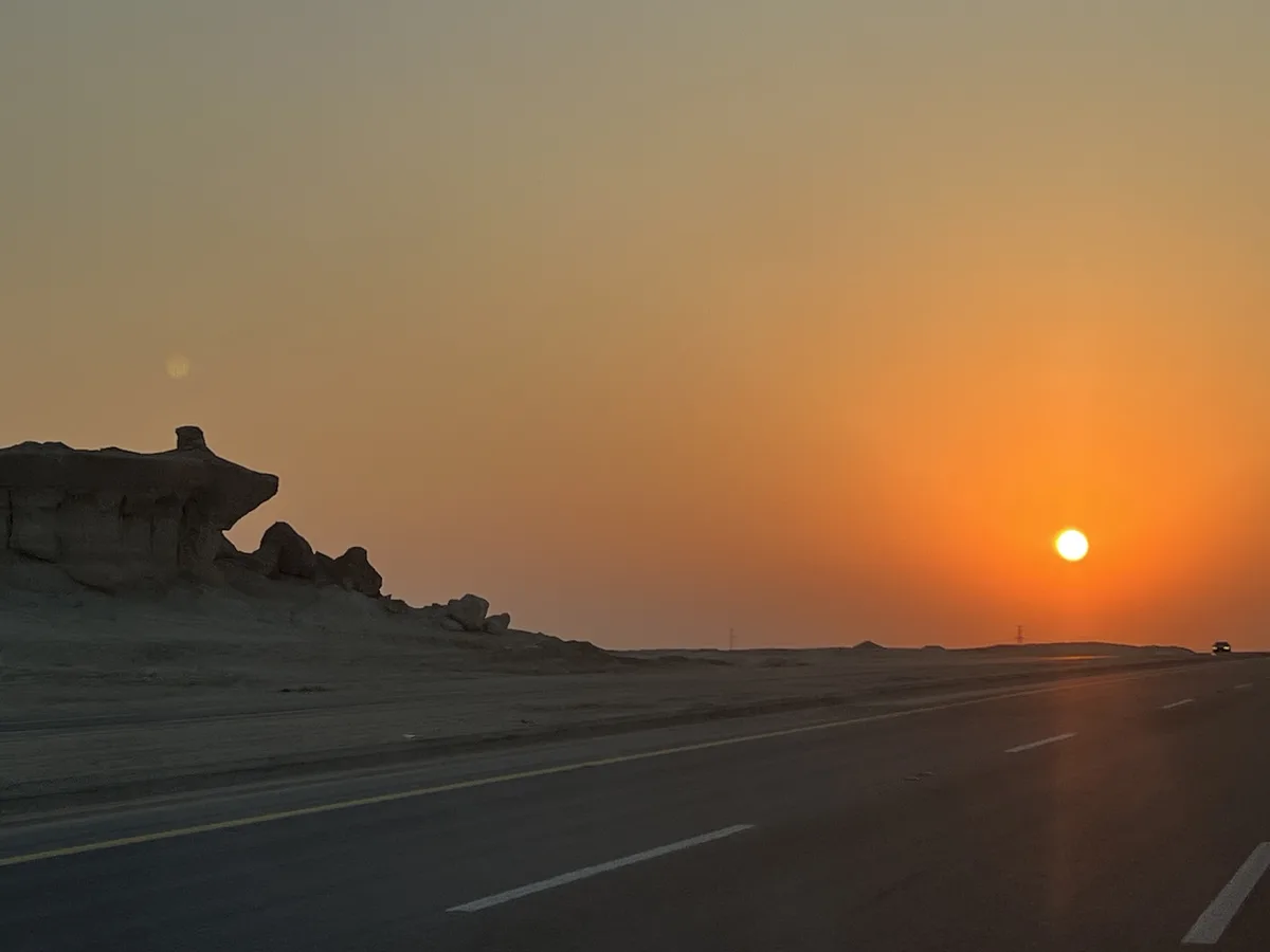 A desert highway at sunset with the sun low on the horizon casting orange light, a large rock formation on the left, and a…