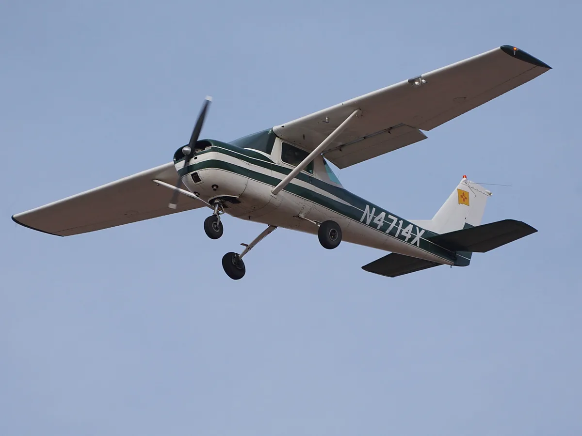 A small white and green single-engine propeller airplane with landing gear down is flying against a clear blue sky. The…