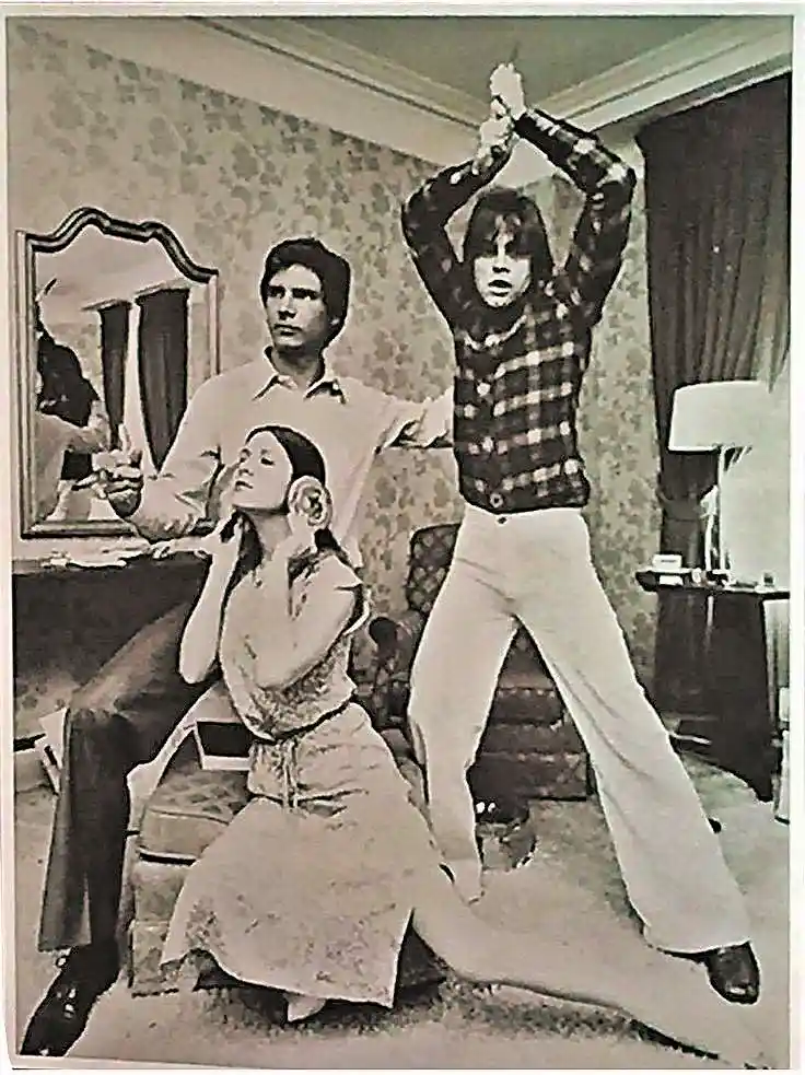 A black-and-white photo shows three people in a 1970s-style room: a man seated holding a glass, a woman seated with her…