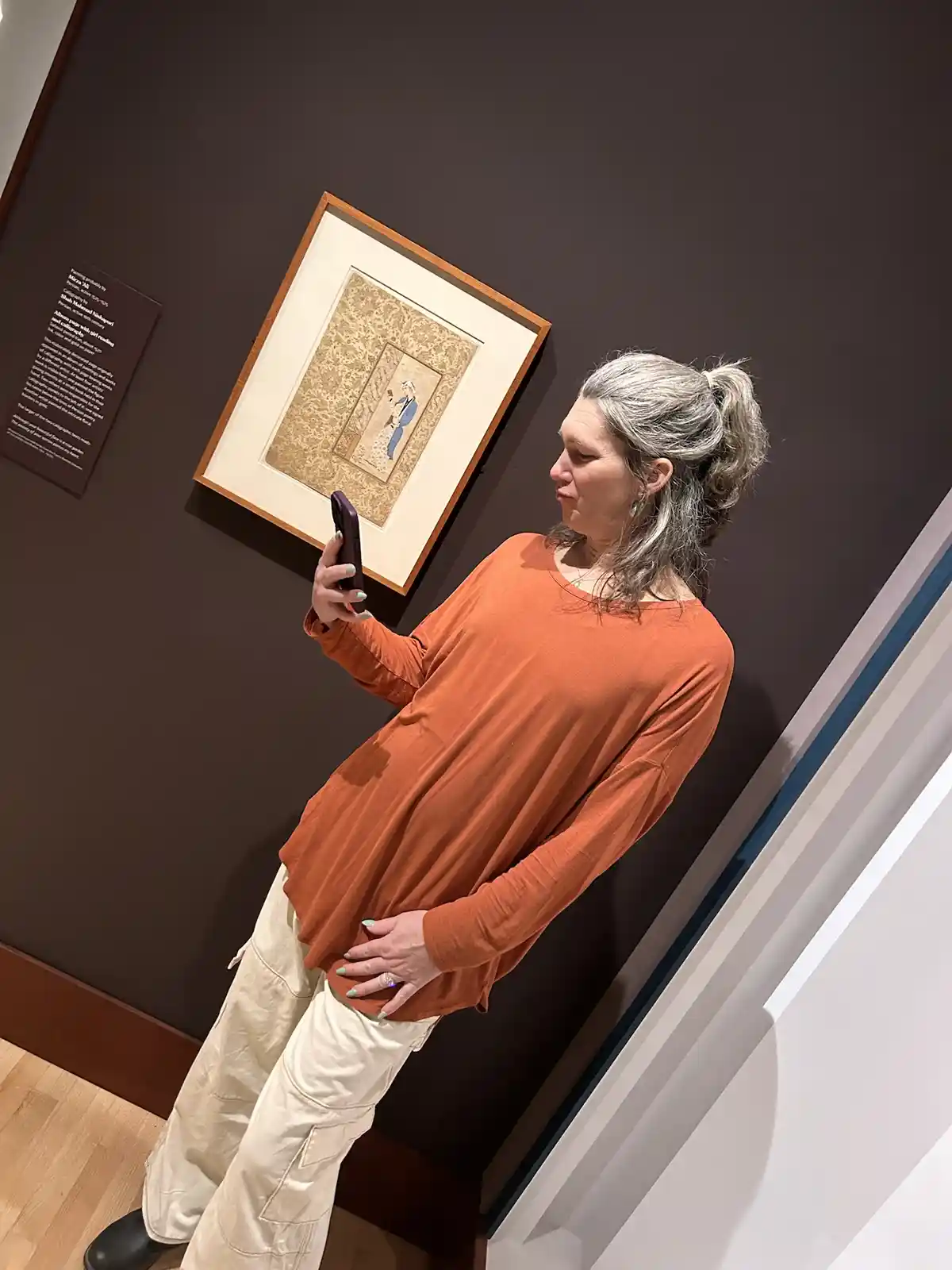 A woman in an orange shirt and beige pants stands in an art gallery, holding her phone up to take a selfie in front of a…