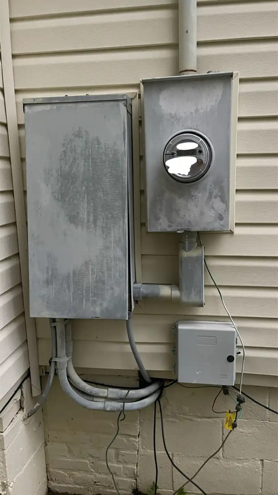 A photo shows two weathered gray utility boxes mounted on beige siding, one housing a circular electric meter with its…