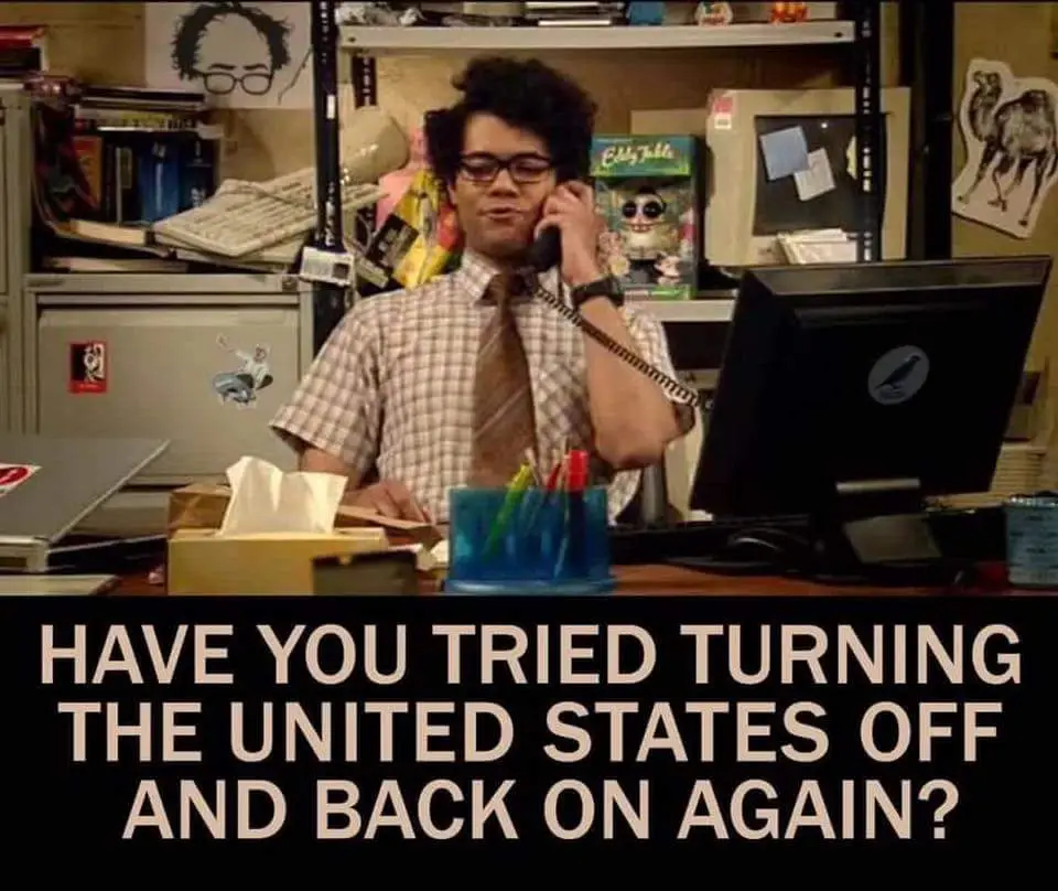 A man with curly hair and glasses sits at a cluttered office desk, holding a phone receiver to his ear while looking…