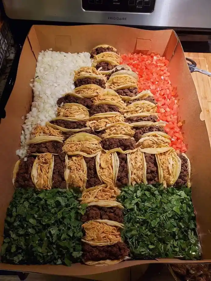 A large cardboard box contains dozens of tacos arranged in rows, flanked by piles of chopped white onions, red tomatoes, and…