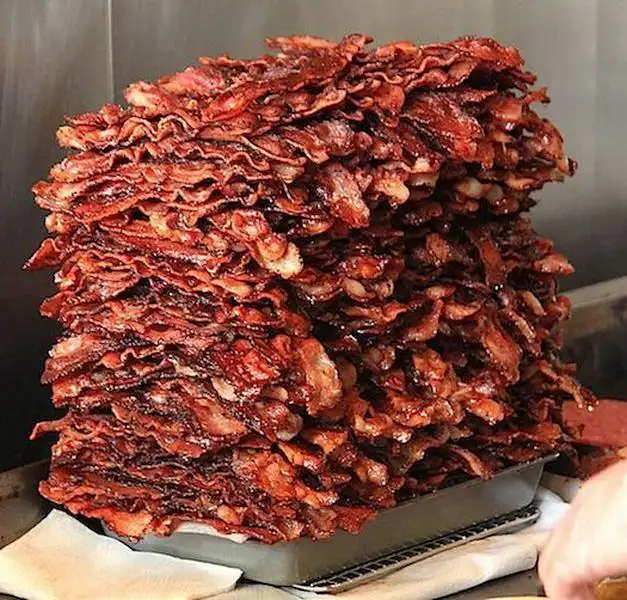 A towering, greasy pile of crispy, cooked bacon strips stacked high in a metal tray, with a hand visible at the bottom…