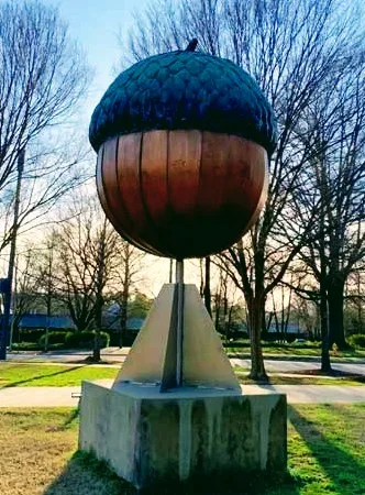 A large, stylized sculpture of an acorn stands on a concrete pedestal in a park with bare trees in the background. The acorn…
