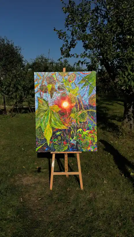 A vibrant, colorful painting on an easel outdoors depicts a sun shining through foliage, with the real sun visible in the…