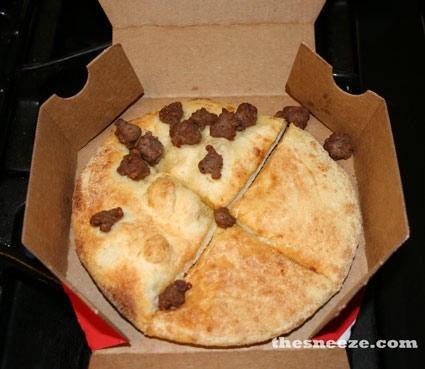 A close-up photo shows a round, golden-brown pizza in a cardboard box, sliced into four pieces, with only a few scattered…