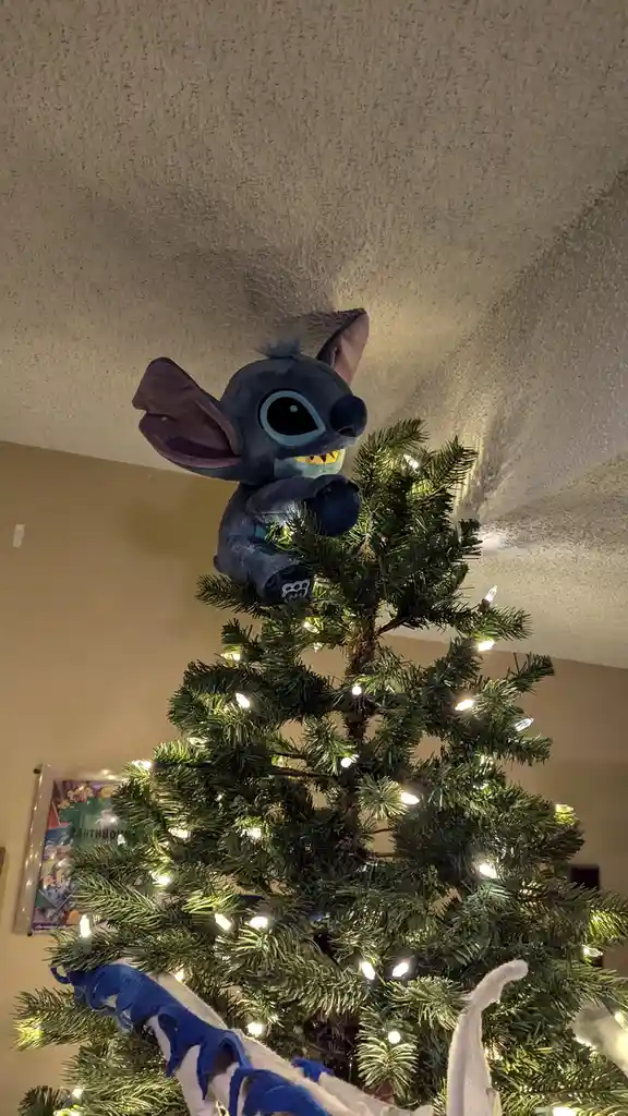 A plush Stitch toy is perched atop a lit Christmas tree, looking down with a wide grin. The tree is decorated with white…