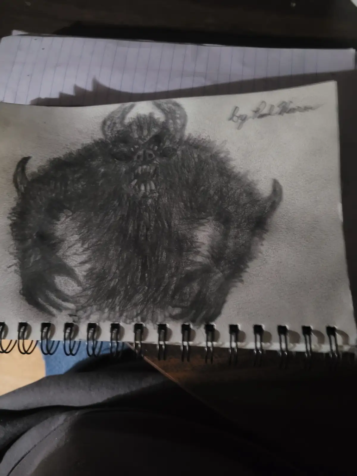 A charcoal sketch on spiral-bound paper depicts a menacing, horned, furry creature with a wide, toothy grin and glowing…