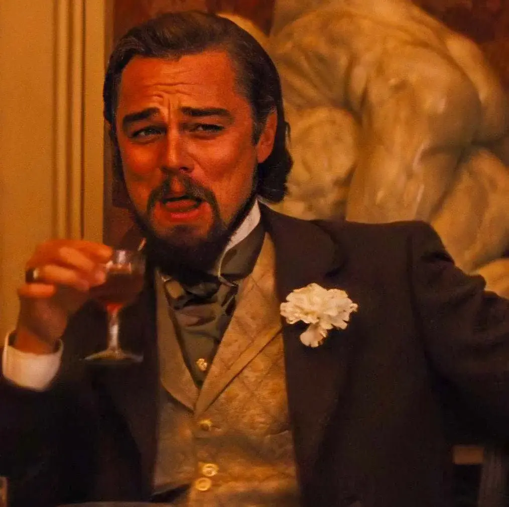 A man in a formal suit with a white flower boutonniere holds a glass of red liquid while looking intensely at the viewer…