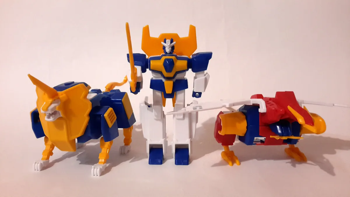 Three plastic toy robots stand side-by-side against a plain white background. The central robot is humanoid, holding a…