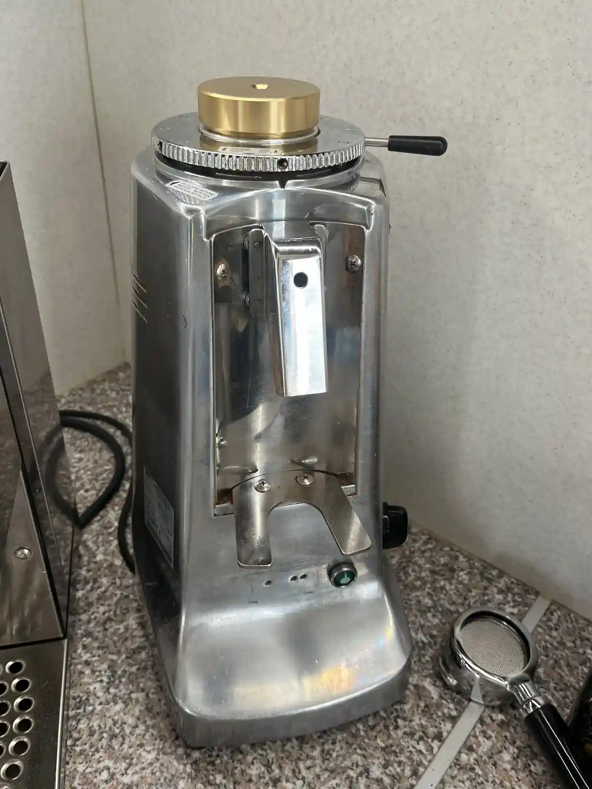 A close-up photo of a silver, industrial-style coffee grinder with a gold top and black handle, sitting on a speckled…
