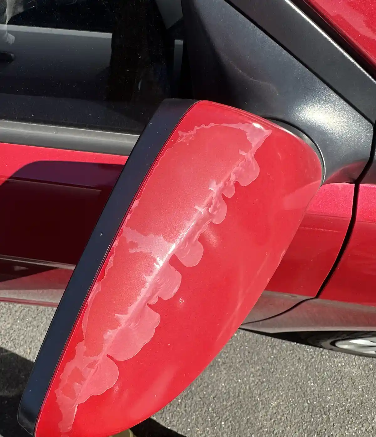A close-up photo shows the side mirror of a red car with a large, irregular patch of clear, glossy substance smeared across…