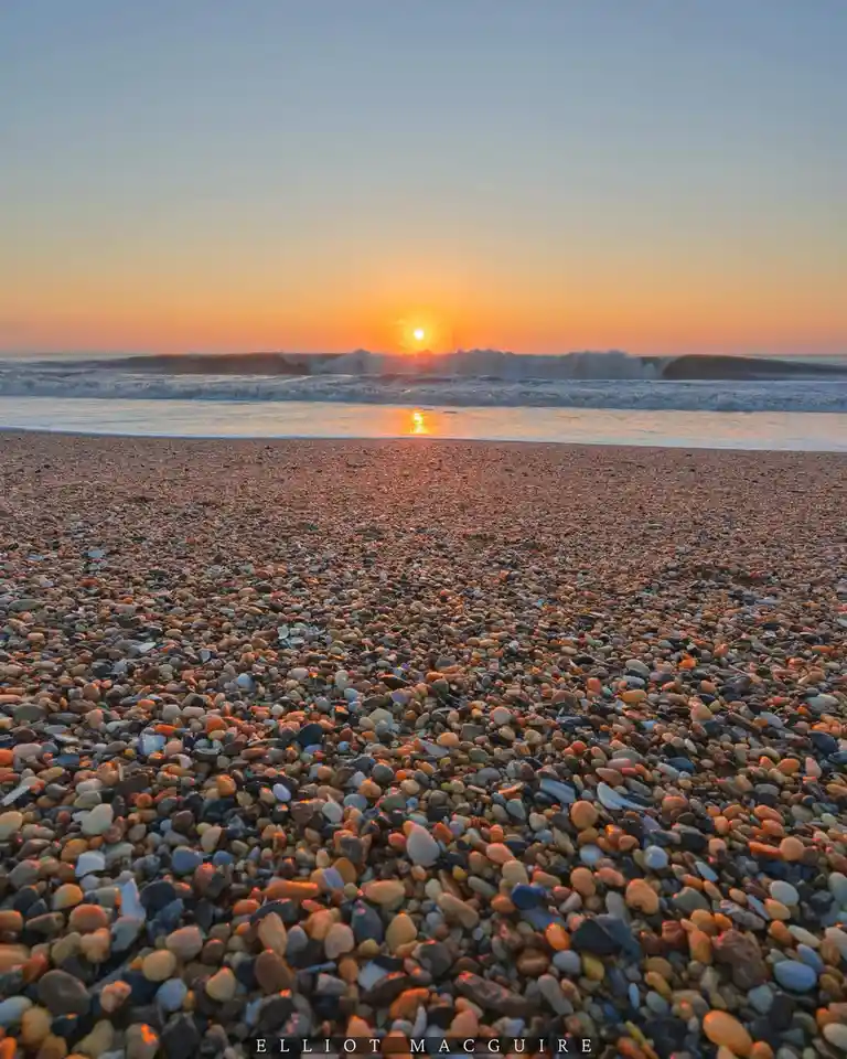 A low-angle photograph of a pebble beach at sunset, with the sun setting over waves in the distance and a watermark reading…