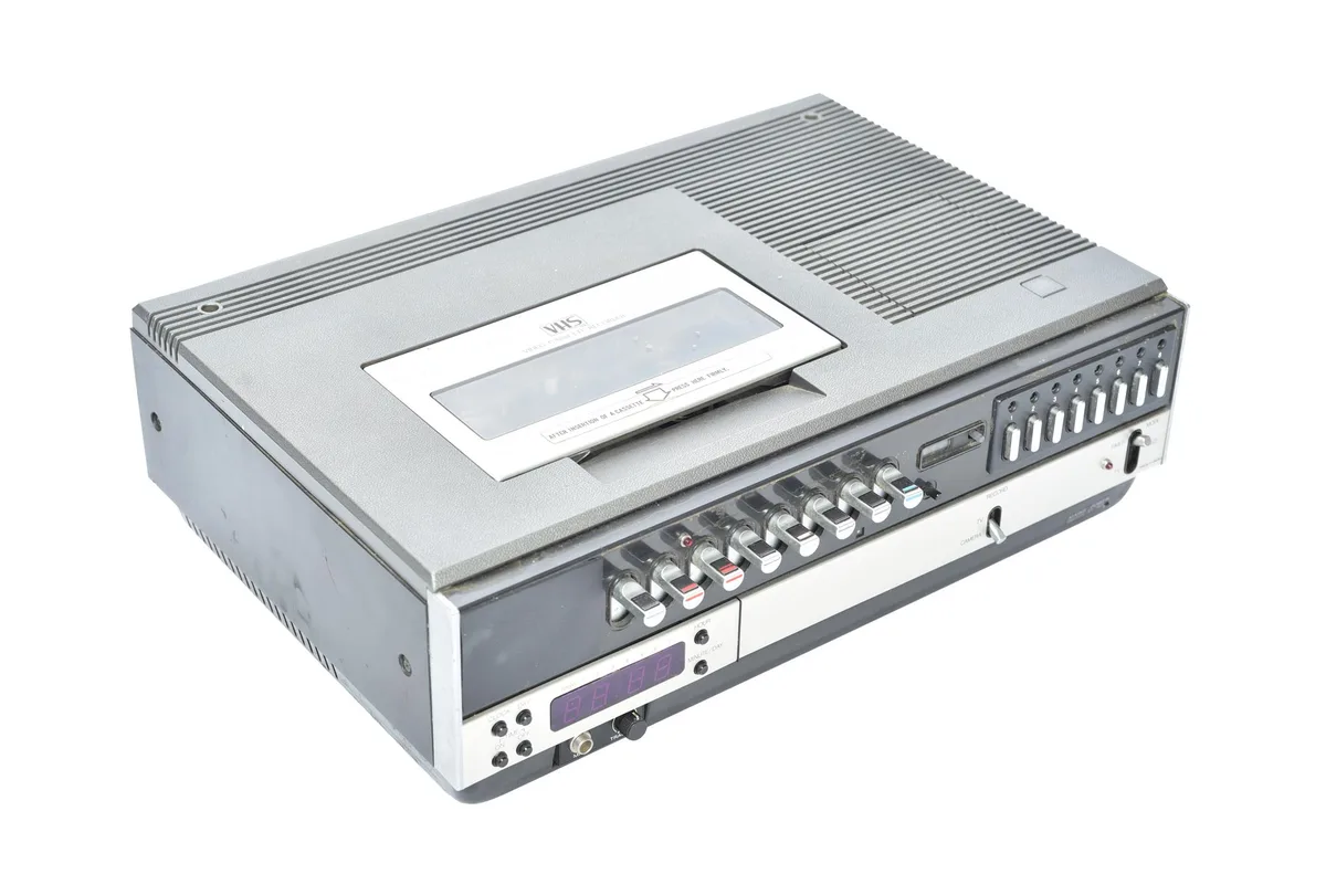 A silver VHS video cassette recorder with a digital display showing '88:88' and numerous knobs and buttons, overlaid with…