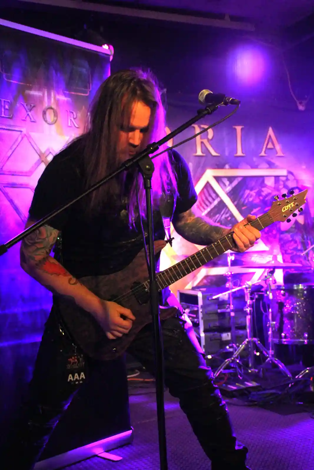 A man with long hair and tattoos plays a Cort electric guitar on stage under purple lights, positioned in front of a…