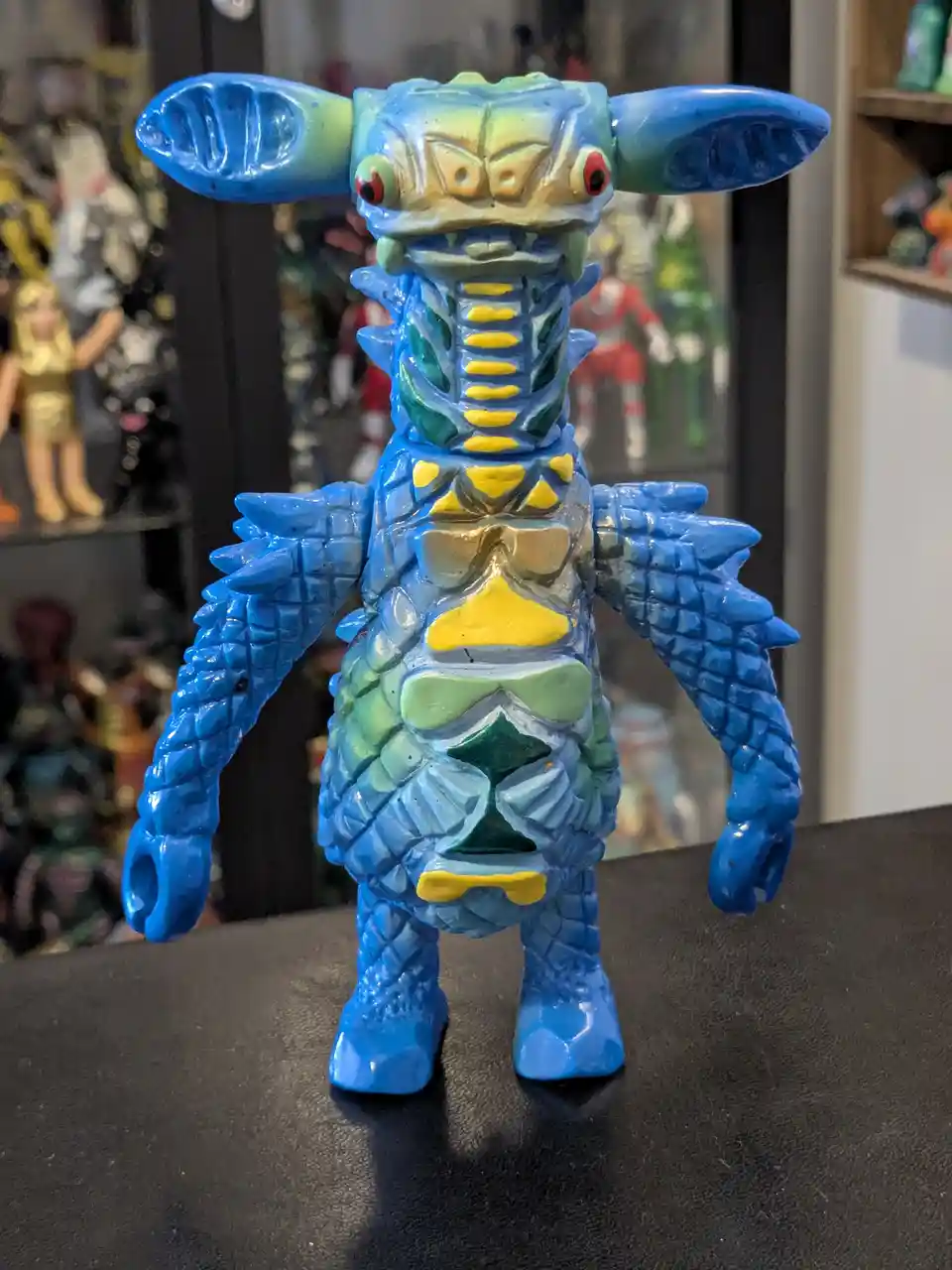A blue, green, and yellow painted monster toy with large ears and a scaly body stands on a black surface, with other toys…