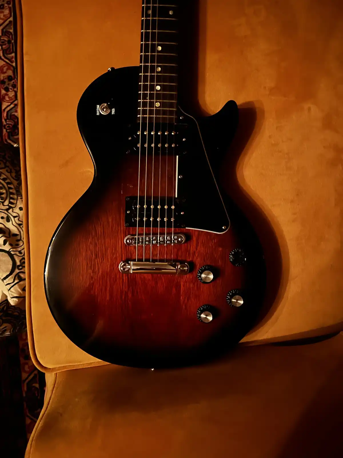A black and sunburst electric guitar with chrome hardware rests on a mustard-yellow velvet couch under warm lighting. The…